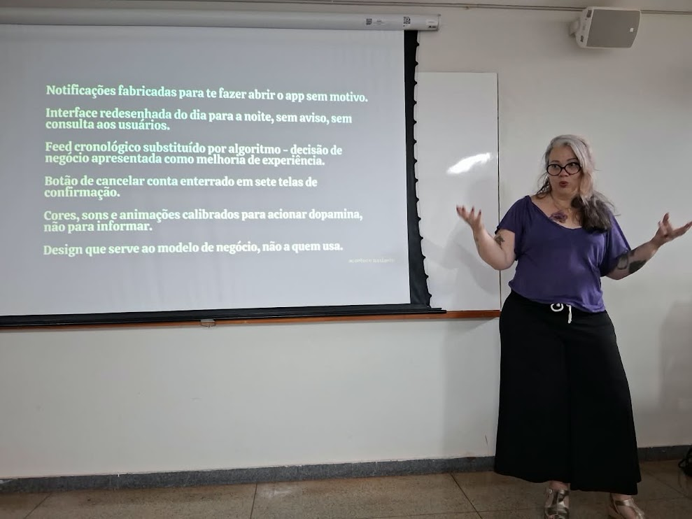 Mulher branca cabelos grisalhos dando palestra sobre Fediverso. Telão exibe motivos para não confiar em redes sociais comerciais.